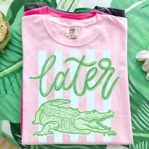 May include: Pink t-shirt with the word "later" in green script, light blue vertical stripes, and a green alligator graphic. The shirt is folded and stacked with other shirts.