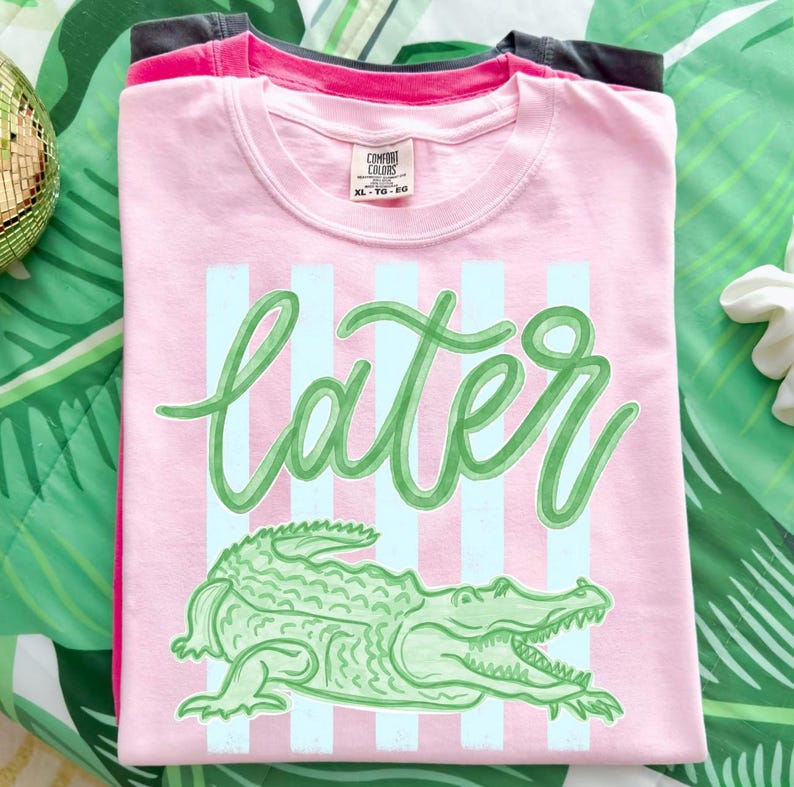 May include: Pink t-shirt with the word "later" in green script, light blue vertical stripes, and a green alligator graphic. The shirt is folded and stacked with other shirts.