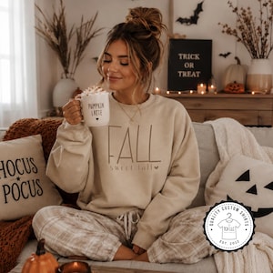 May include: A beige crewneck sweatshirt featuring the text FALL and sweet fall with a small heart icon. The garment is paired with light beige plaid lounge pants. A white mug held in the image displays the text PUMPKIN LATTE with a heart icon.