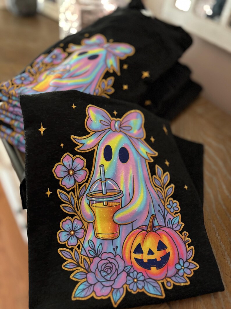 May include: A black t-shirt featuring a graphic of a holographic-style ghost wearing a bow, holding an iced beverage with a straw, and standing next to a jack-o'-lantern and purple flowers. The design includes small gold star accents.