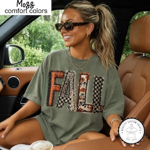 May include: A moss green short-sleeve t-shirt featuring the word FALL in large block letters across the chest. Each letter has a different pattern including orange plaid, leopard print, floral, and black and white checkerboard with lace trim.