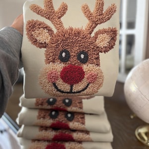 May include: A cream-colored sweatshirt featuring a textured, three-dimensional punch needle embroidery of a reindeer face. The reindeer has brown fur, large antlers, black eyes, and a prominent red nose, with a stack of identical sweatshirts below.