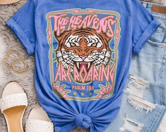 May include: A blue short-sleeve t-shirt featuring a graphic print of a tiger face with the text "THE HEAVENS ARE ROARING" and "PSALM 19:1" in pink and white, surrounded by a light green border and pink lightning bolt accents.