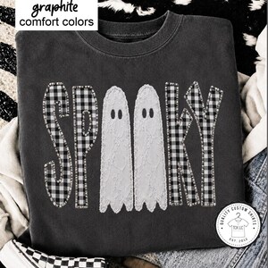 May include: A graphite-colored crewneck sweatshirt featuring the text SPOOKY in a black and white buffalo plaid pattern, with two white lace ghost silhouettes replacing the letter O. The text graphite comfort colors is visible at the top left.