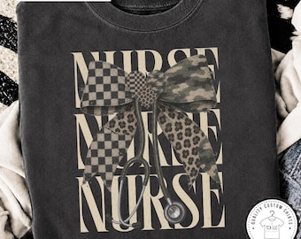 Pode incluir: Uma camisola de gola redonda cor de grafite com o texto "NURSE" repetido numa fonte grande e clara. Ao centro, um gráfico de um estetoscópio com um laço grande com padrões axadrezados, de leopardo e de camuflagem na frente.
