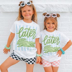 May include: Two young girls wearing matching white t-shirts with the word "later" in green script and a green alligator graphic. The shirts have blue vertical stripes. One girl wears black and white checkered shorts, the other wears pink and white checkered shorts.