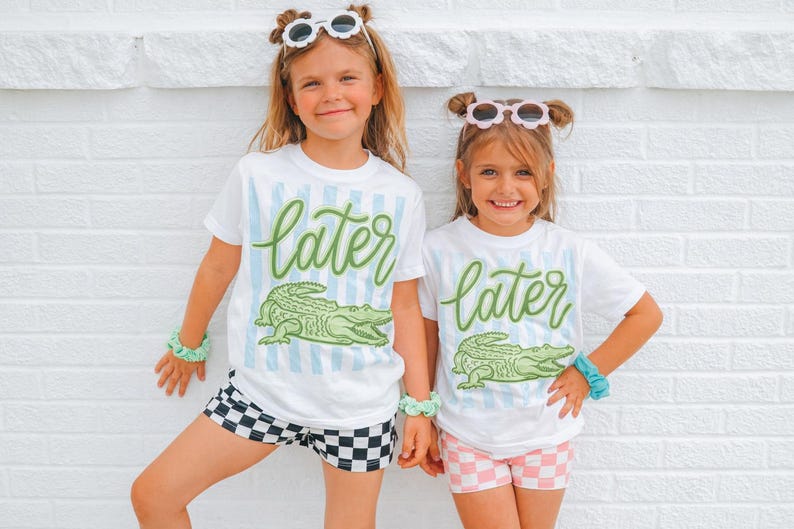 May include: Two young girls wearing matching white t-shirts with the word "later" in green script and a green alligator graphic. The shirts have blue vertical stripes. One girl wears black and white checkered shorts, the other wears pink and white checkered shorts.