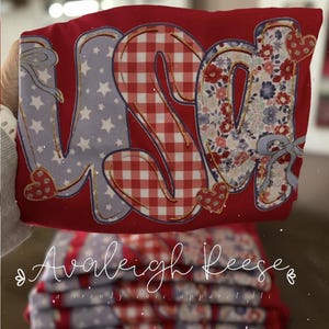 May include: Red t-shirt with the word "USA" in a patchwork design. The letters feature patterns of stars, gingham, and floral prints. The shirt is folded, with the brand name "Avalleigh Reese" and "a trendy, chic, apparel llc" visible.