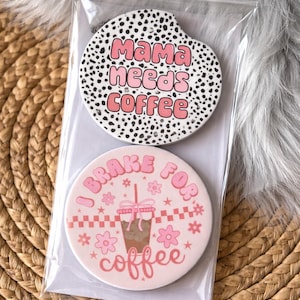 May include: Two round car coasters in a clear plastic sleeve. One coaster has a black and white dalmatian print with the text "Mama needs coffee" in pink. The other coaster is pink with a coffee cup graphic and the text "I brake for coffee".