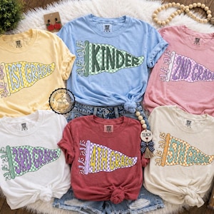 May include: A collection of six short-sleeved t-shirts in various colors, each featuring a pennant design with a grade level printed on it. The shirts are in yellow, blue, pink, white, red, and cream. The pennants have a checkered pattern.