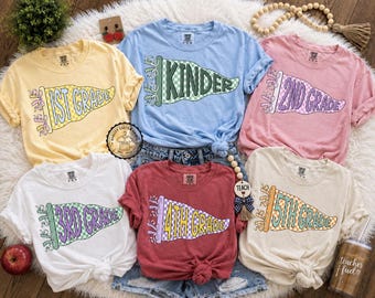 May include: A collection of six short-sleeved t-shirts in various colors, each featuring a pennant design with a grade level printed on it. The shirts are in yellow, blue, pink, white, red, and cream. The pennants have a checkered pattern.