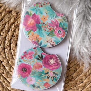 May include: Two floral car coasters. Each coaster features a teal background with pink, orange, and white flowers. The coasters are packaged in a clear plastic sleeve and rest on a woven wicker surface.