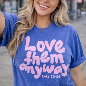 May include: Blue t-shirt with the text "Love them anyway" in pink, with "Luke 23:34" below. The shirt has rolled-up sleeves and is worn with a dark blue cap and a gold cross necklace.