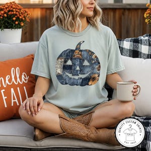 May include: A light sage green short-sleeve t-shirt featuring a large graphic of a jack-o'-lantern pumpkin composed of blue denim patchwork patterns and orange sunflower accents. The shirt has a crew neckline and a relaxed, loose-fitting cut.