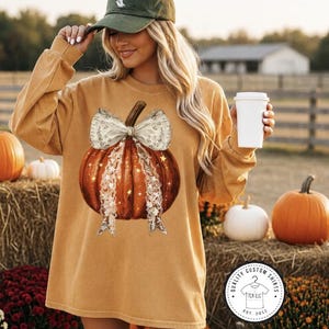 May include: A mustard yellow long-sleeve sweatshirt featuring a graphic of an orange pumpkin adorned with a large white lace bow and small star accents. The sweatshirt has a crew neckline and a relaxed fit, showcasing a seasonal autumn design.