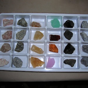May include: A white plastic rectangular tray with twenty-four individual compartments, each containing a unique mineral specimen. The collection includes various rocks and crystals in shades of pink, gray, yellow, green, brown, black, and orange.