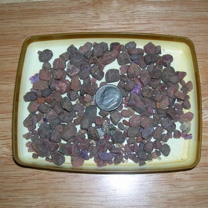 May include: A collection of small, irregularly shaped, dark reddish-brown stones fills a rectangular, light yellow tray with a gold rim. A silver dime is placed among the stones, providing a size reference. The stones appear to be minerals or gemstones.