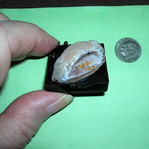 May include: A small, open geode with a light tan exterior and a white interior, containing gold-colored flecks, sits on a black base. A U.S. dime is next to the geode for size comparison. The background is a light green color.