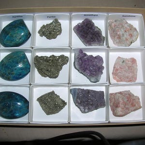 May include: A collection of twelve mineral specimens organized in a cardboard box with white dividers. The set includes three polished blue apatite stones, three metallic pyrite clusters, three purple amethyst crystals, and three peach-colored sunstone pieces.