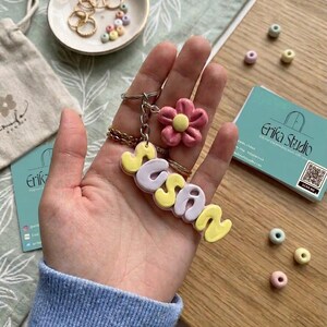 May include: A handmade keychain spelling "Susan" in yellow and lilac letters, with a pink flower charm and gold chain. The keychain is held in a hand, with a business card and small dish of jewellery in the background.
