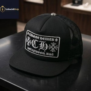 Puede incluir: Gorra de camionero negra con parte trasera de malla y visera curva. La parte delantera presenta un parche rectangular blanco con el texto "CHROME HEARTS" y "CH HOLLYWOOD, USA" en una fuente estilizada. Un botón de metal plateado en la parte superior.