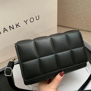 May include: A black rectangular shoulder bag featuring a quilted grid pattern with padded square sections. The bag includes a thin adjustable black strap with a silver-toned metal buckle and a flap closure. The background shows a bag with the text HANK YOU.