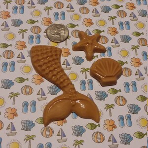 May include: A set of three brown resin summer-themed decorations. The decorations include a mermaid tail, a seashell, and a starfish. The decorations are on a white background with a repeating pattern of suns, palm trees, flip-flops, sailboats, fish, and flowers.