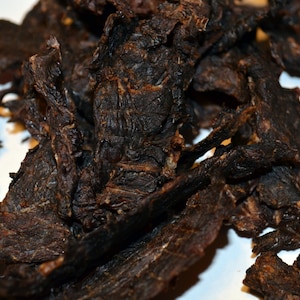 May include: A close-up of a pile of dark brown beef jerky. The jerky is dried and has a rough texture.