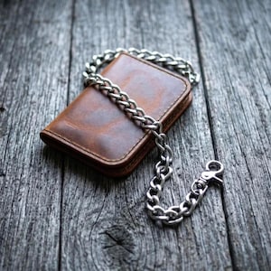 Puede incluir: Una cartera rectangular de cuero marrón con un acabado liso y desgastado y esquinas redondeadas. Una cadena gruesa de metal plateado pulido está unida a la cartera, con un cierre de mosquetón resistente en el extremo para su sujeción.
