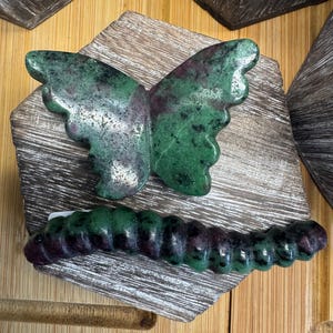 May include: A green and maroon stone butterfly and caterpillar. The butterfly, with detailed wings, sits above the caterpillar. Both are presented on a wooden surface, showcasing the natural stone colours.