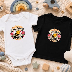 May include: A white short-sleeved bodysuit and a black short-sleeved t-shirt, both featuring a central graphic of a smiling bee surrounded by pink flowers and the text "Don't Worry HAPPY" in a circular, sunburst-inspired design.