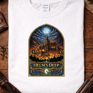 May include: White t-shirt featuring a detailed graphic of a castle under siege, with flames and a full moon. The design includes the text "Helm's Deep" in a decorative banner. The shirt is made of a soft material.