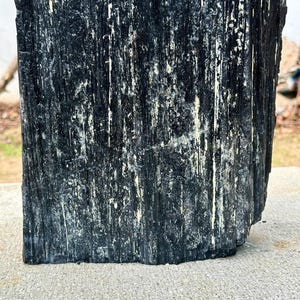 Puede incluir: Un esp&eacute;cimen de cristal de turmalina negra en bruto que presenta una estructura columnar profundamente estriada con una textura rugosa y dentada. La superficie muestra surcos verticales y crestas con sutiles toques de inclusiones minerales claras.