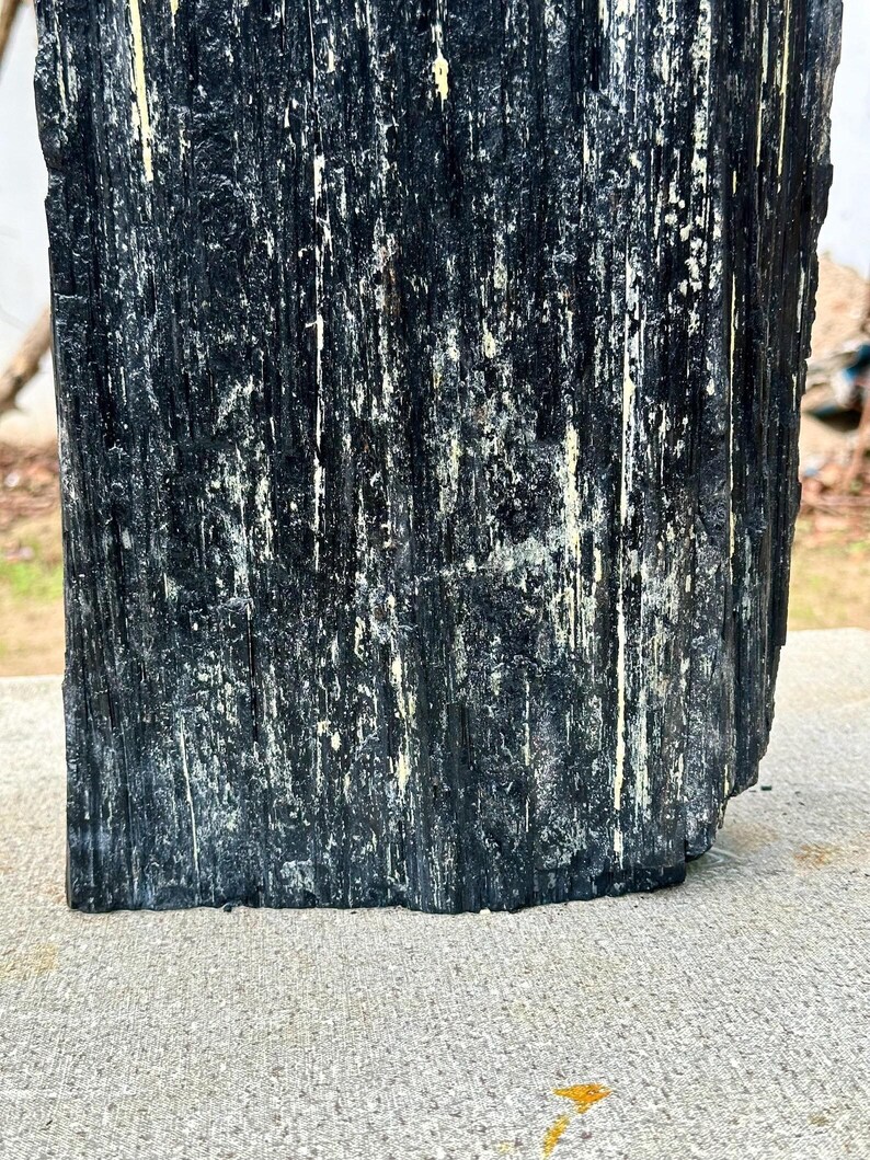 Puede incluir: Un esp&eacute;cimen de cristal de turmalina negra en bruto que presenta una estructura columnar profundamente estriada con una textura rugosa y dentada. La superficie muestra surcos verticales y crestas con sutiles toques de inclusiones minerales claras.