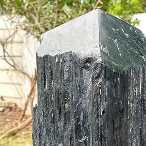 Puede incluir: Un esp&eacute;cimen de cristal de turmalina negra de gran altura con una textura rugosa y estriada verticalmente en los lados y una terminaci&oacute;n piramidal plana, lisa y pulida en el &aacute;pice, que muestra formaciones minerales naturales y tonos oscuros.
