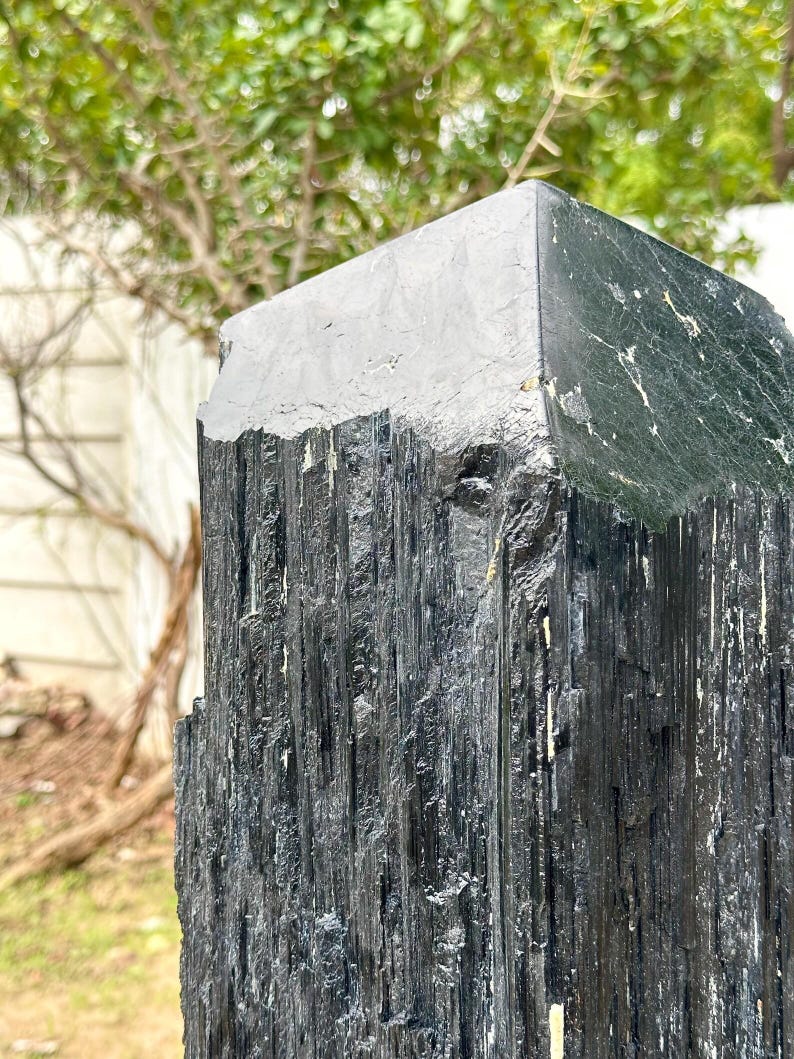 Puede incluir: Un esp&eacute;cimen de cristal de turmalina negra de gran altura con una textura rugosa y estriada verticalmente en los lados y una terminaci&oacute;n piramidal plana, lisa y pulida en el &aacute;pice, que muestra formaciones minerales naturales y tonos oscuros.
