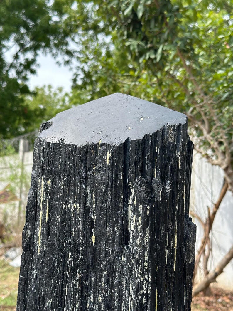 Puede incluir: Un esp&eacute;cimen de cristal de turmalina negra de gran altura con una superficie superior hexagonal plana y lados verticales profundamente estriados con una apariencia rugosa y texturizada. El mineral muestra un color oscuro y opaco con inclusiones.
