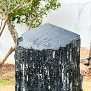 Puede incluir: Un esp&eacute;cimen de cristal de turmalina negra, alto y vertical, con una superficie superior plana y hexagonal. El exterior presenta estr&iacute;as longitudinales paralelas y profundas a lo largo de su cuerpo, con sutiles inclusiones minerales claras.