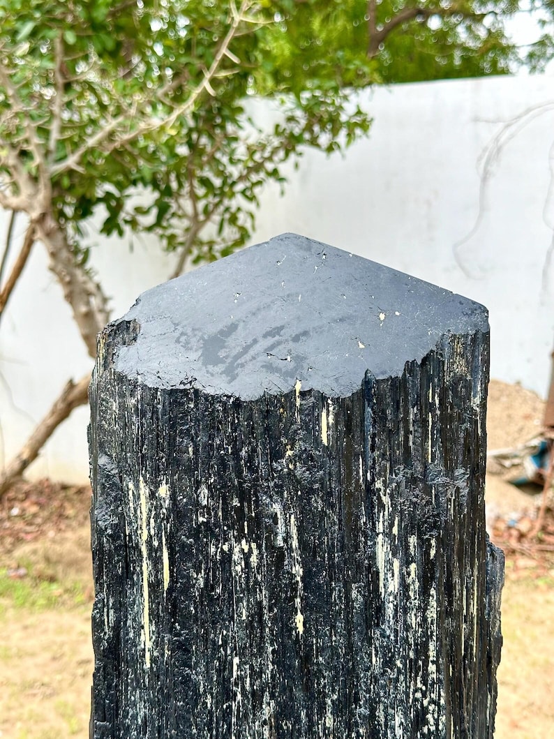 Puede incluir: Un esp&eacute;cimen de cristal de turmalina negra, alto y vertical, con una superficie superior plana y hexagonal. El exterior presenta estr&iacute;as longitudinales paralelas y profundas a lo largo de su cuerpo, con sutiles inclusiones minerales claras.