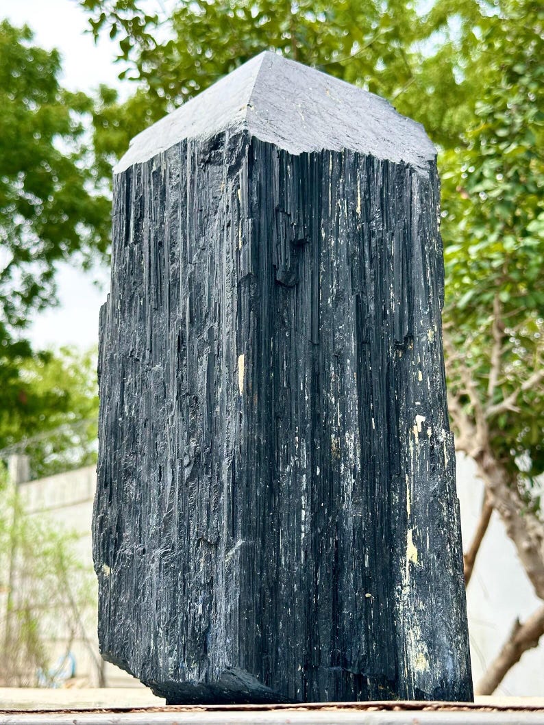 Puede incluir: Una punta de cristal de turmalina negra alta con una textura rugosa de estr&iacute;as verticales y una parte superior naturalmente facetada. La piedra presenta una coloraci&oacute;n negro carb&oacute;n con sutiles inclusiones minerales claras.