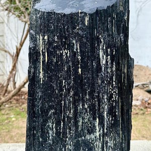 Puede incluir: Un esp&eacute;cimen alto de cristal de turmalina negra en bruto con una estructura columnar estriada verticalmente y una superficie superior plana y pulida. El mineral oscuro y opaco presenta un color negro profundo con sutiles inclusiones.
