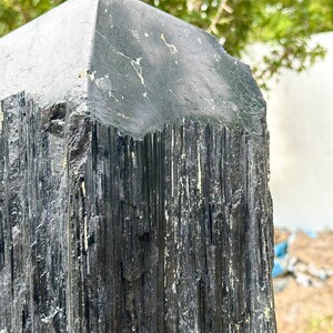 Puede incluir: Un esp&eacute;cimen de cristal de turmalina negra con una punta de terminaci&oacute;n naturalmente facetada. La superficie muestra estr&iacute;as verticales profundas y una apariencia rugosa y texturizada con sutiles inclusiones minerales blancas.