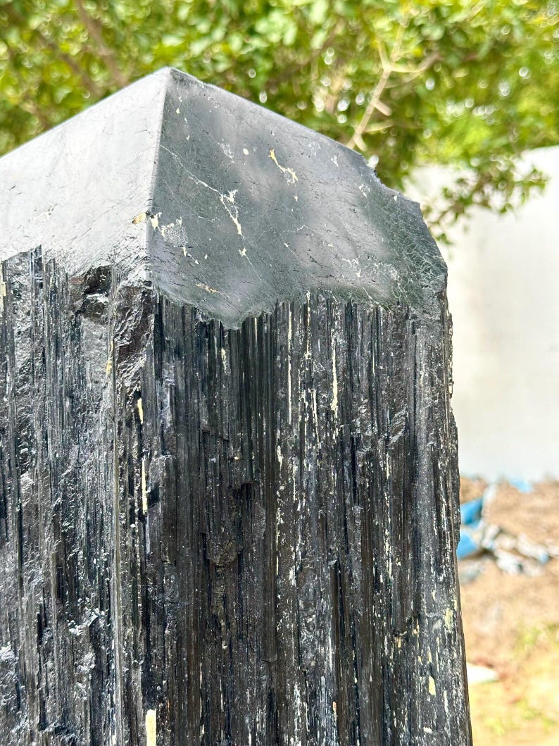 Puede incluir: Un esp&eacute;cimen de cristal de turmalina negra con una punta de terminaci&oacute;n naturalmente facetada. La superficie muestra estr&iacute;as verticales profundas y una apariencia rugosa y texturizada con sutiles inclusiones minerales blancas.