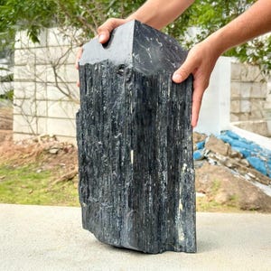 Puede incluir: Un gran esp&eacute;cimen de cristal de turmalina negra en bruto con una estructura columnar estriada verticalmente y una superficie oscura, brillante y opaca. El mineral presenta bordes afilados y una textura natural sin pulir con surcos.