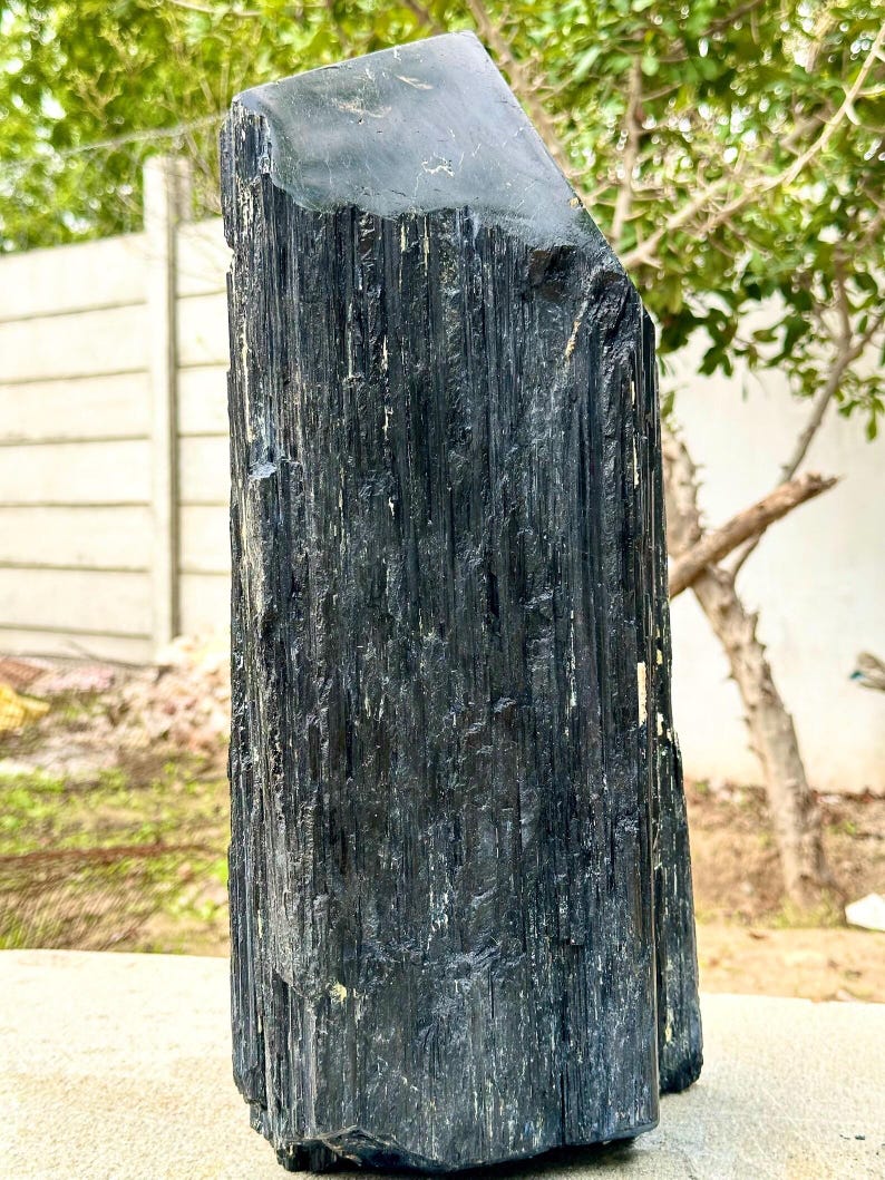 Puede incluir: Un esp&eacute;cimen de cristal de turmalina negra en bruto, alto, con una estructura columnar profundamente estriada y una superficie superior lisa e inclinada. El mineral presenta un aspecto oscuro y opaco con surcos verticales.