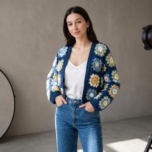 May include: A woman wearing a navy blue crochet cardigan with floral patterns in yellow, white, and light blue. The cardigan is open-front and paired with a white top and blue jeans. The background is a neutral color.