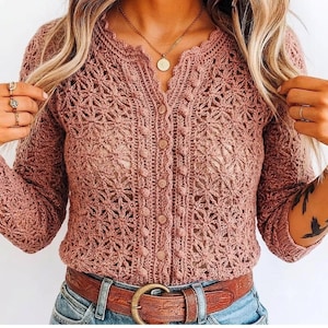 Peut inclure: Un cardigan à manches longues en crochet rose poudré présentant un motif floral délicat, une encolure festonnée et une rangée verticale de boutons assortis au centre. Le vêtement est porté avec une ceinture en cuir marron et un jean bleu.