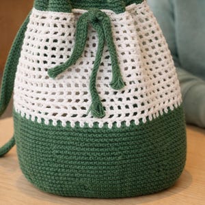 Peut inclure: Un sac à cordon fait à la main en crochet blanc et vert. Le sac présente une base et une bordure supérieure vertes unies, avec un corps blanc en tissage ouvert. Une fermeture à cordon et une sangle vertes complètent le design. Le sac est posé sur une surface en bois.