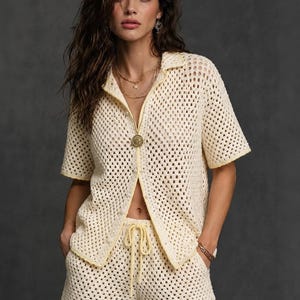Puede incluir: Un conjunto de dos piezas de ganchillo en color crema que consta de una camisa de manga corta con botones y cuello, y pantalones cortos a juego con cintura de cordón. La prenda presenta un patrón de punto abierto con ribete amarillo claro.