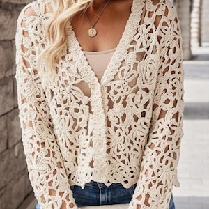 May include: A cream-colored long-sleeve cardigan featuring an intricate openwork crochet pattern with floral motifs. The garment has a V-neckline, a front button closure, and long sleeves. It is worn over a solid light-colored camisole and blue jeans.